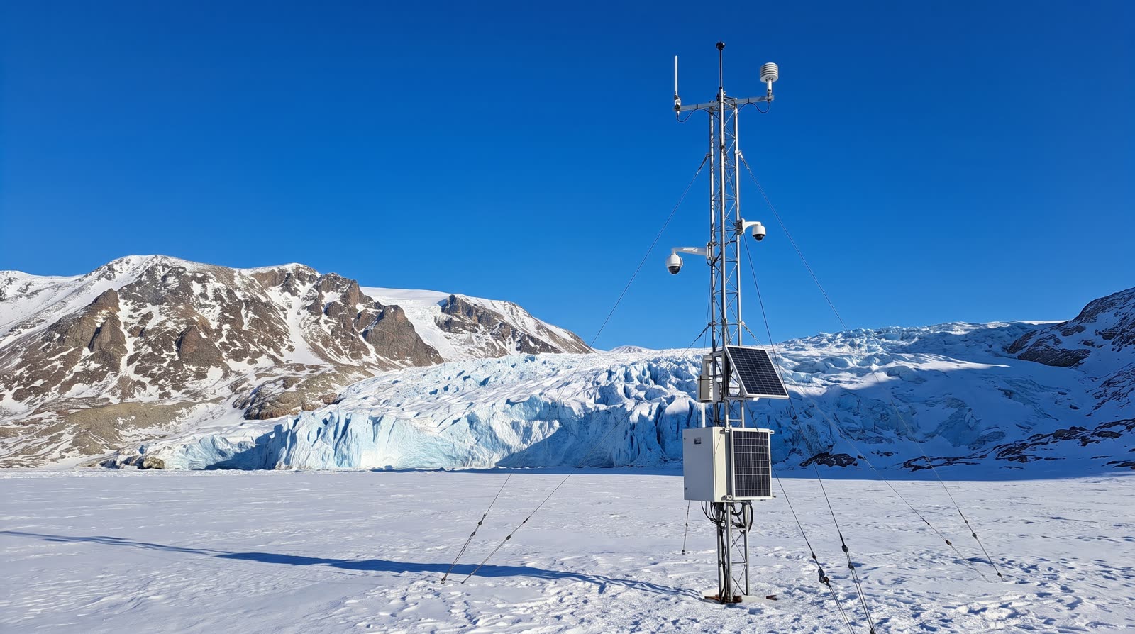 A weather mast with solar panels on a snowfield below a glacier front, under a deep blue sky