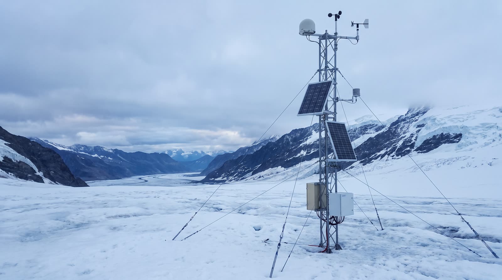 A weather and GNSS mast with solar panels standing on a glacier that runs down a mountain valley