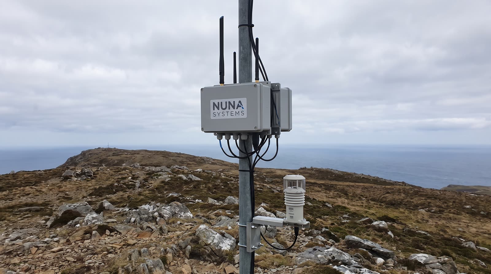 A grey NUNA node clamped to a mast beside a weather sensor, on a rocky headland above the sea