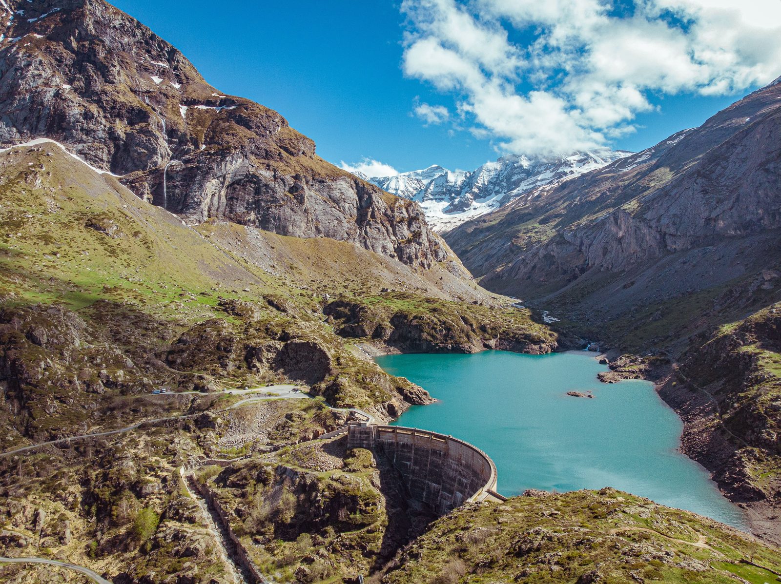 An arch dam holding a turquoise reservoir in a steep mountain valley, snowy peaks behind