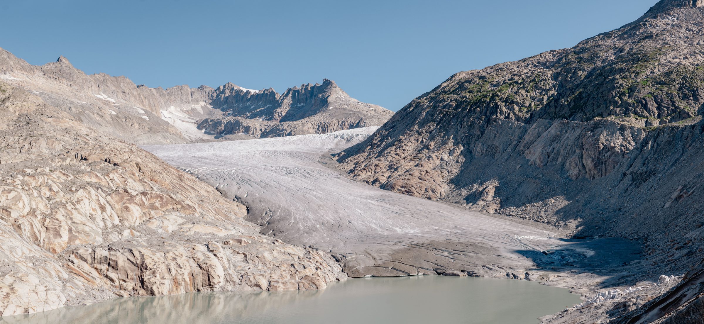 A valley glacier flowing between bare grey mountains into a meltwater lake, under a clear sky