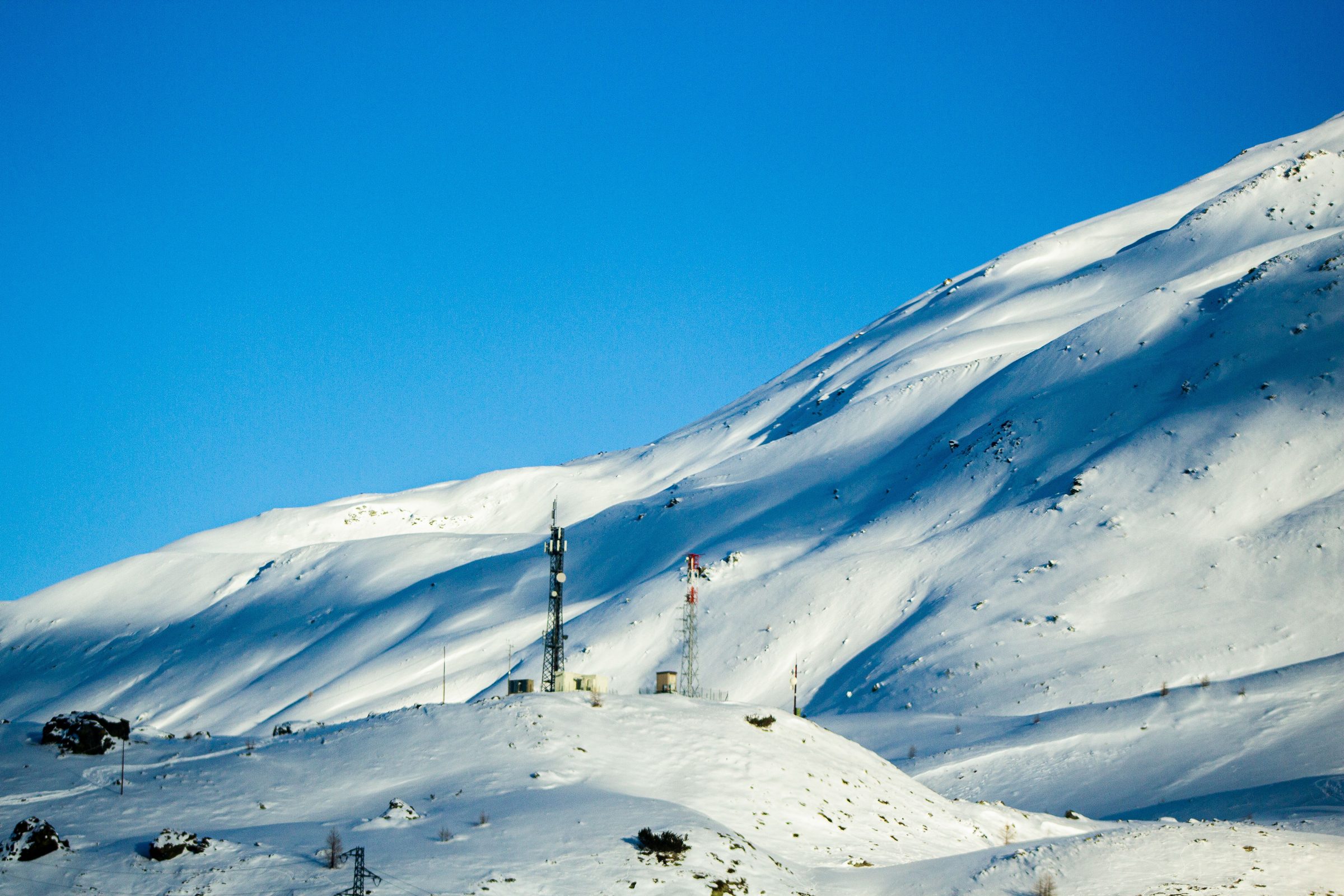 Radio masts and small equipment huts on a snow-covered mountain shoulder under a deep blue sky