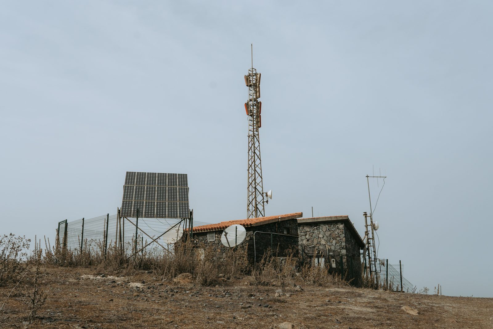 A small stone equipment hut with a lattice radio mast, satellite dishes and a bank of solar panels on a dry hilltop