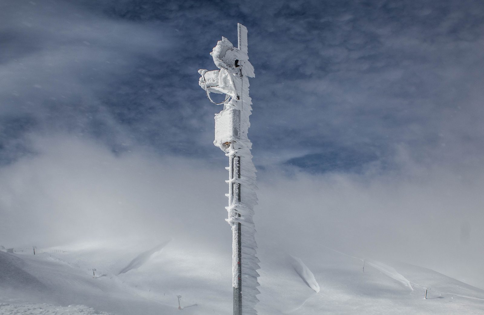 An instrument mast coated in thick rime ice, standing in deep snow under a cloudy blue sky