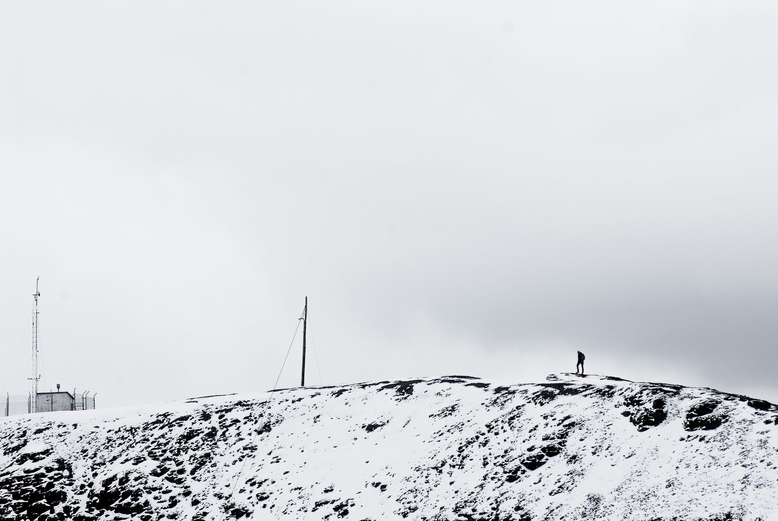 A lone person standing on a snowy, rocky ridge beside instrument masts, under a white overcast sky
