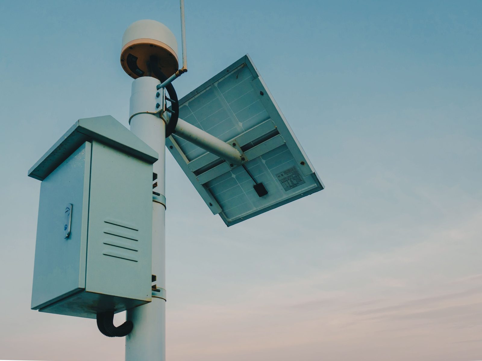 A logger box, a small solar panel and a GNSS antenna on an instrument pole against a pale sky