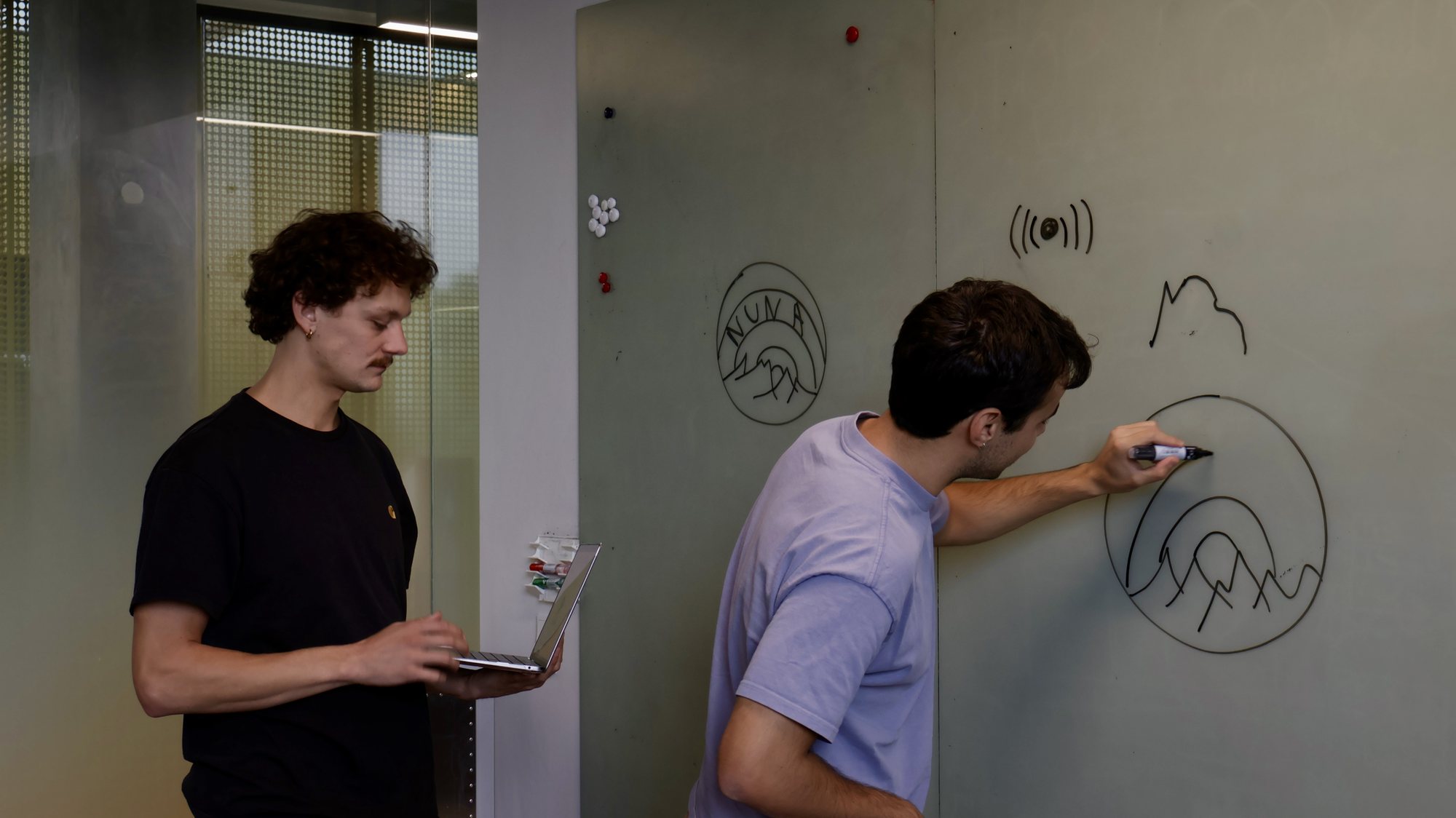 Nikitas drawing mountains and the NUNA logo on a whiteboard while Rafael works on a laptop beside him