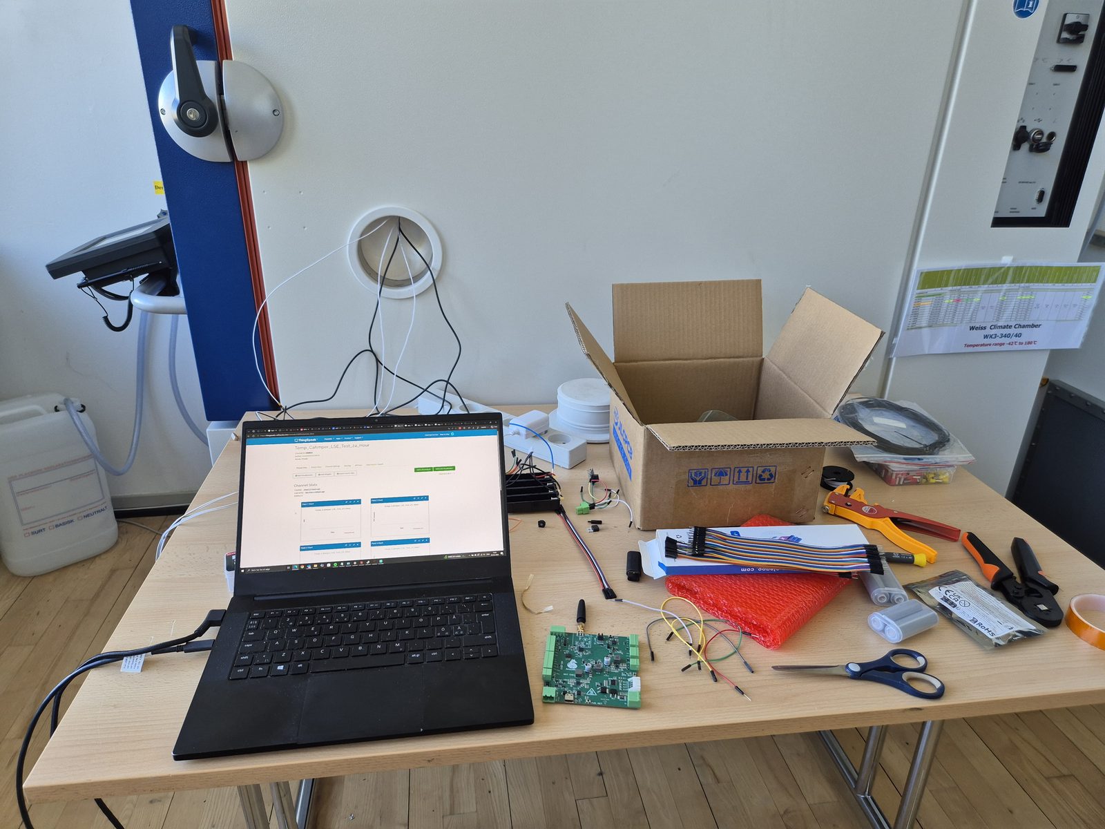 Our test desk beside the climate chamber at DTU: a V1 board, a laptop showing the live test data, cables and tools, with sensor leads running through the chamber wall